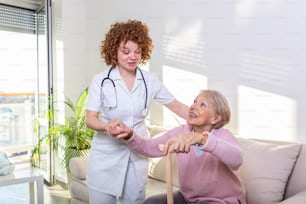 Female caregiver helping senior woman get up from couch in living room. Smiling nurse assisting senior woman to get up. Caring nurse supporting patient while getting up from sofa