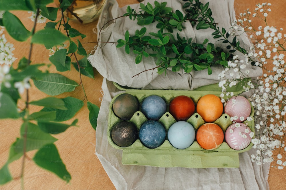 Natural dye easter eggs in carton tray on rustic table with flowers. Modern yellow, pink, blue and grey easter eggs painted with organic onion, beets, red cabbage, carcade tea. Zero waste holiday