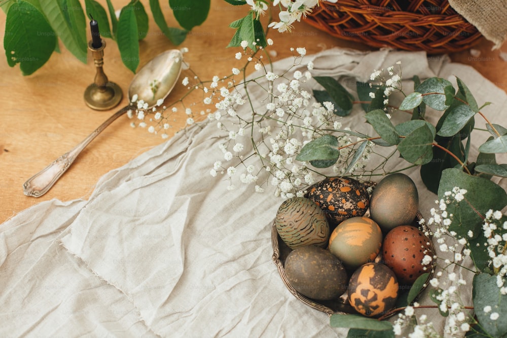 촛불과 바구니가 있는 소박한 나무 테이블에 봄 꽃과 유칼립투스가 있는 현대 부활절 달걀. 세련된 회색 돌과 녹색 부활절 달걀은 카케이드 차의 천연 염료로 칠해져 있습니다.