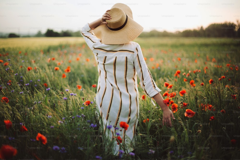Junge Frau in rustikalem Leinenkleid spaziert zwischen Mohn und Kornblumen in sommerlicher Wiesenlandschaft im Sonnenuntergangslicht. Ländliches langsames Leben. Das einfache Leben genießen. Mädchen mit Hut in Wildblumen