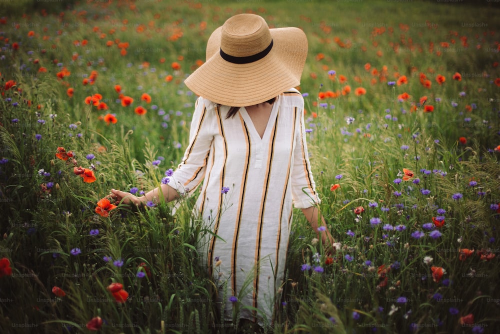 Young woman in linen dress and hat enjoying rural evening among poppy and cornflowers in countryside. Stylish girl in rustic dress walking in wildflowers in warm light in summer meadow.