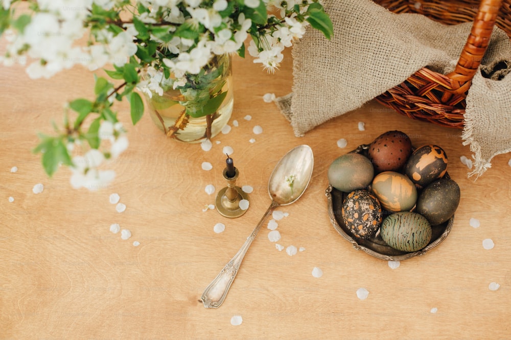 행복한 부활절 플랫 레이. 바구니가 있는 소박한 나무 테이블에 봄 꽃이 있는 현대 부활절 달걀. 세련된 회색 돌과 녹색 부활절 달걀은 카케이드 차의 천연 염료로 칠해져 있습니다.