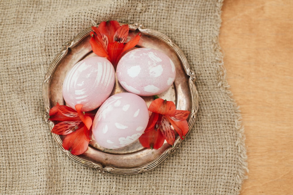 Ovos de Páscoa modernos com flores vermelhas no prato vintage na mesa de madeira rústica, flat lay. Elegantes ovos de Páscoa rosa pastel pintados em corante natural de beterraba. Feliz Páscoa