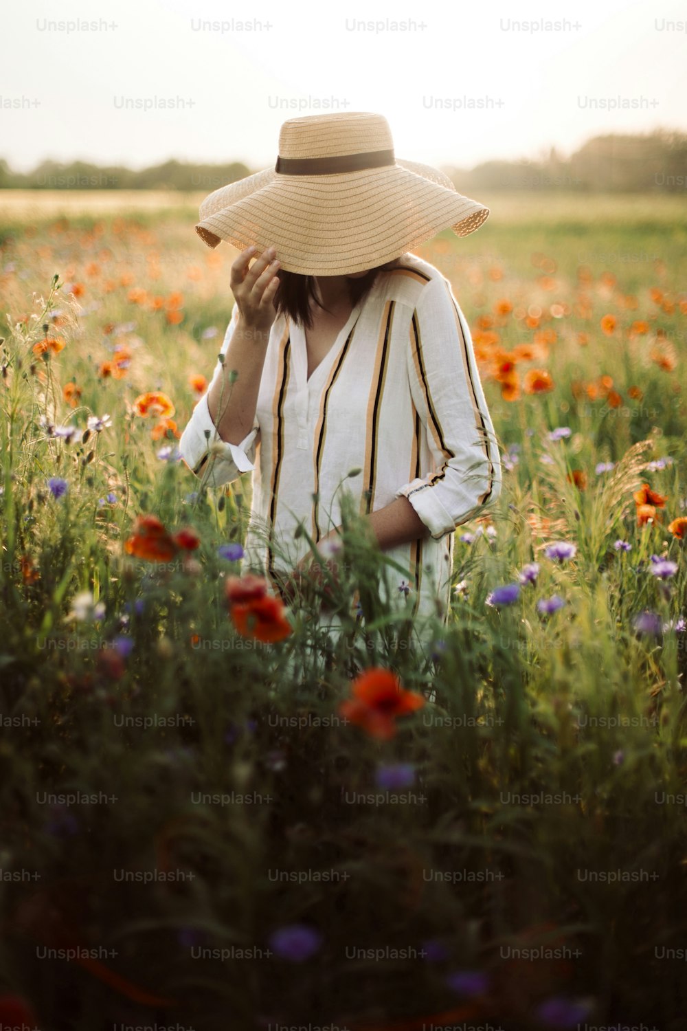 Young woman in rustic linen dress walking among poppy and cornflowers in summer meadow countryside in sunset light. Rural slow life. Enjoying simple life. Girl in hat in wildflowers
