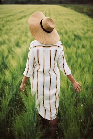 Young woman in linen dress and hat walking in green field of barley in countryside. Stylish girl in rustic dress enjoying peaceful moment in grass in summer. Tranquil rural moment