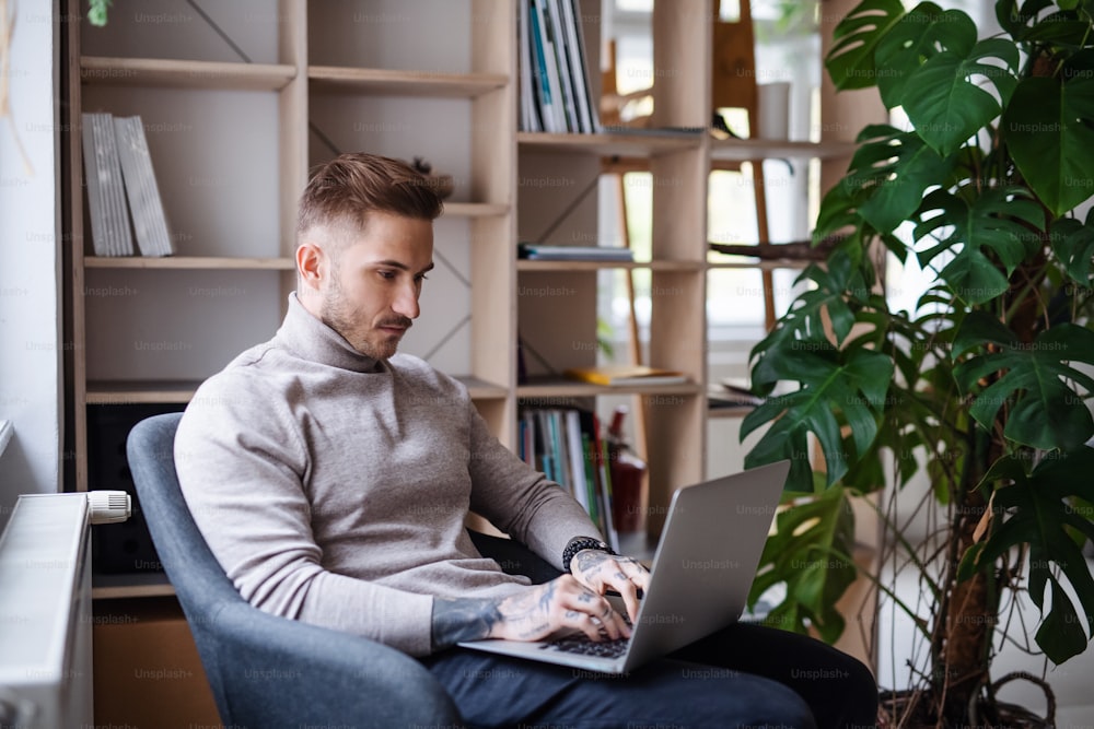 Um jovem empresário sentado em poltrona dentro de casa no escritório, usando laptop.