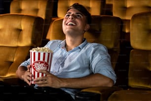A young man smiling enjoying with his popcorn while watching a movie in theater cinema