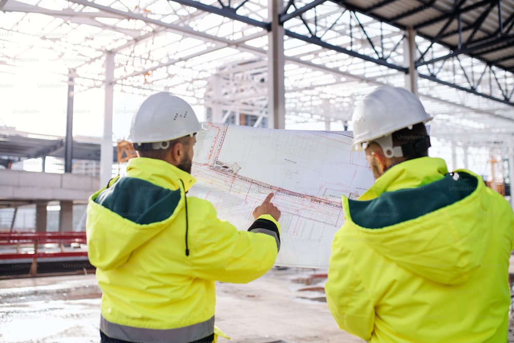 Rear view of men engineers standing outdoors on construction site, holding blueprints.