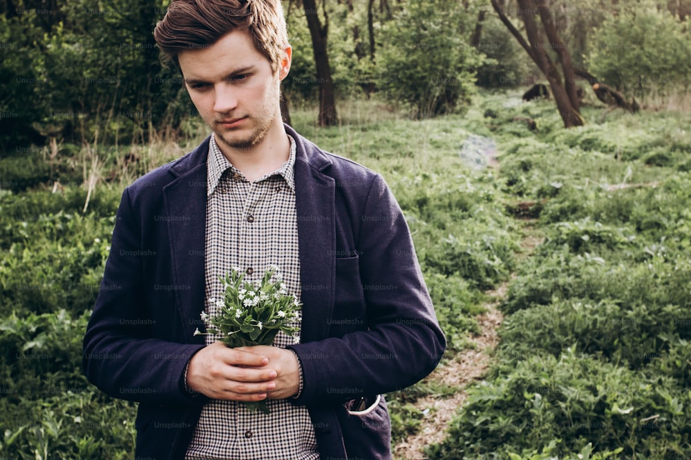 stylish hipster man holding spring bouquet of wildflowers in sunny countryside and waiting. space for text. rustic groom with simple flowers in hands. beautiful moment. save environment.