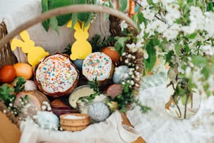 Oeufs modernes de Pâques, pain de Pâques, décorations de lapin, jambon, betteraves, beurre, saucisses dans un panier rustique décoré de branches de buis vert et de fleurs sur une table en bois avec bougie