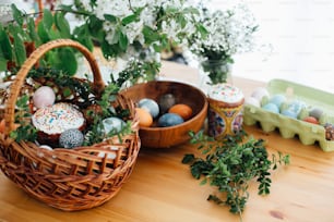 Ovos de Páscoa modernos, pão de Páscoa, presunto, beterraba, manteiga, em cesto de vime decorado com ramos de buxus verdes e flores sobre mesa de madeira rústica. Cesta de Páscoa tradicional.
