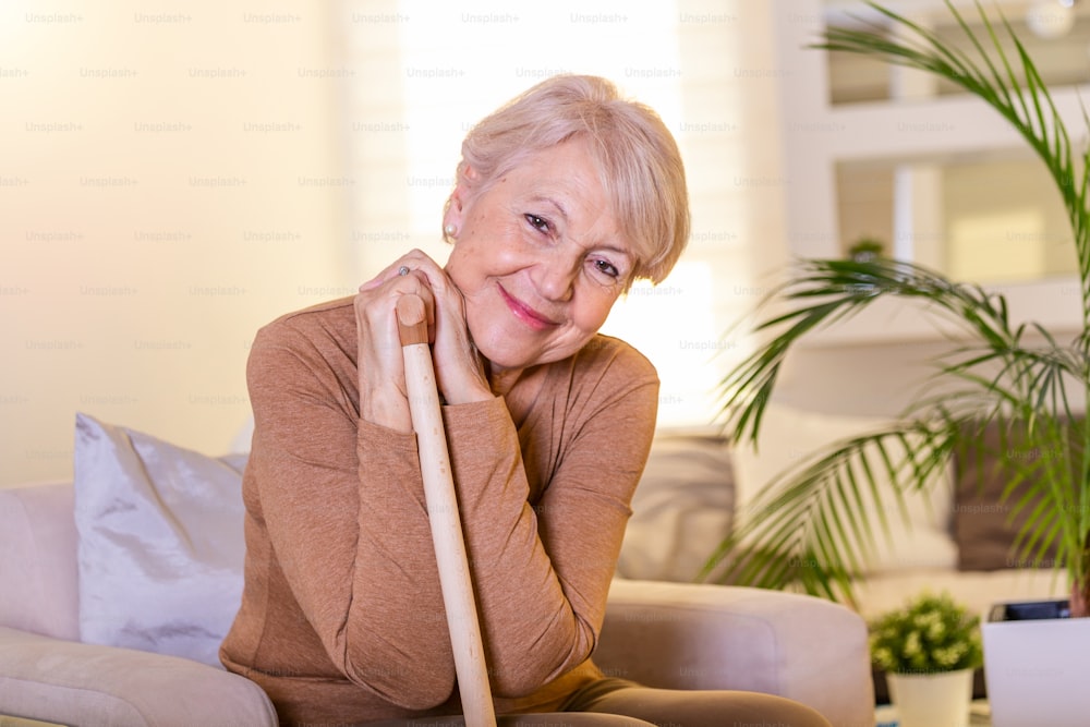 Pleasant thoughtful aged woman seating with a walking stick. Retired woman with her wooden walking stick at home. Happy senior woman relaxing at home holding cane and looking at camera.