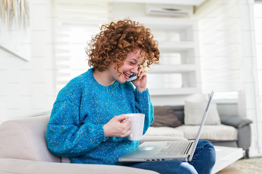 Young businesswoman sitting at her desk with laptop talking on mobile phone. Young red hair beautiful woman drinking coffee, using smartphone and laptop, smiling and looking at laptop
