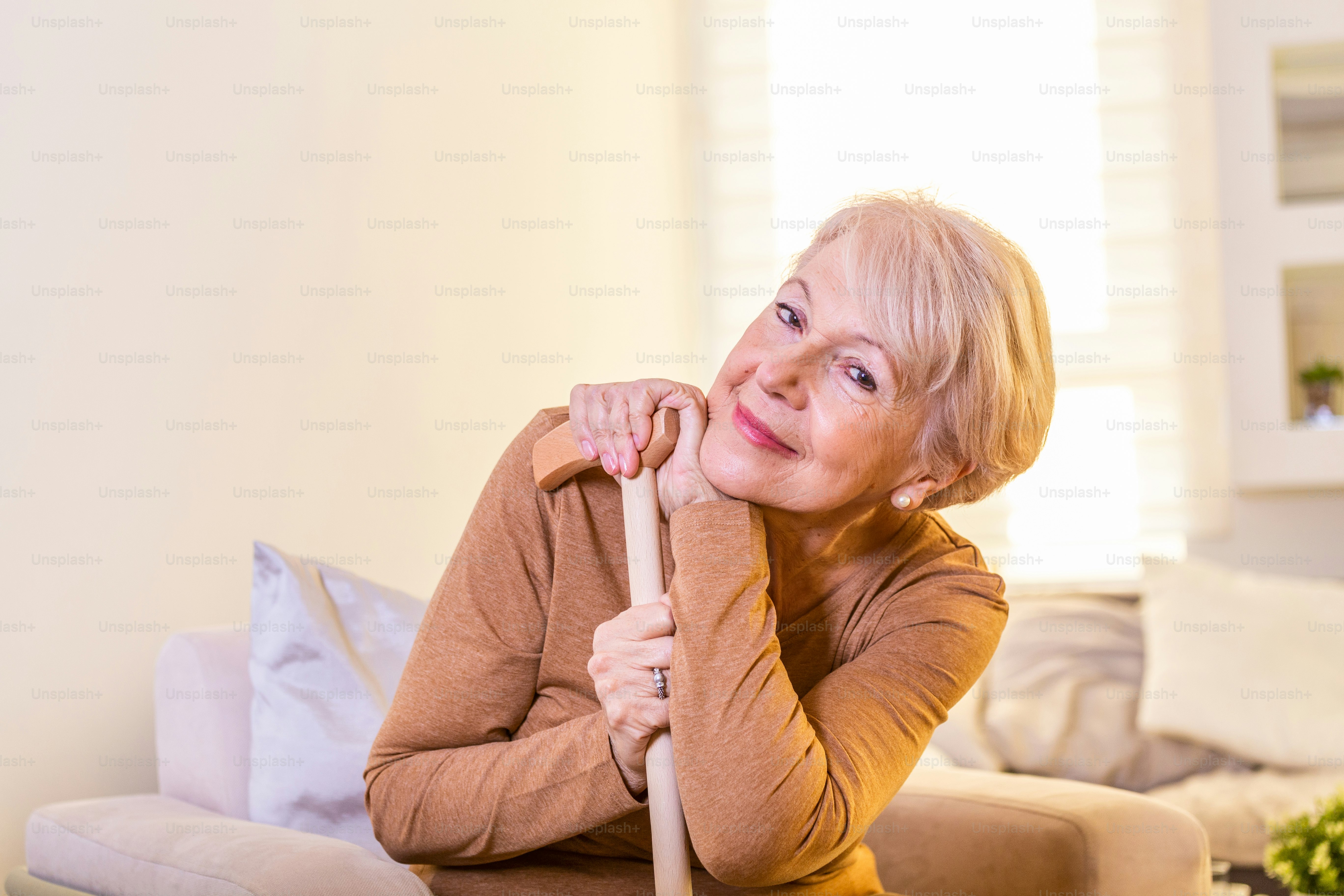 Smiling grandmother sitting on couch. Portrait of a beautiful smiling senior woman with walking cane on light background at home. Old woman sitting with her hands on a cane