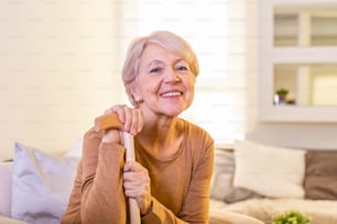 Porträt einer schönen älteren Frau mit weißen Haaren und Gehstock. Porträt einer älteren Frau, die zu Hause auf dem Sofa sitzt. Lächelnde reife grauhaarige Frau mittleren Alters schaut in die Kamera