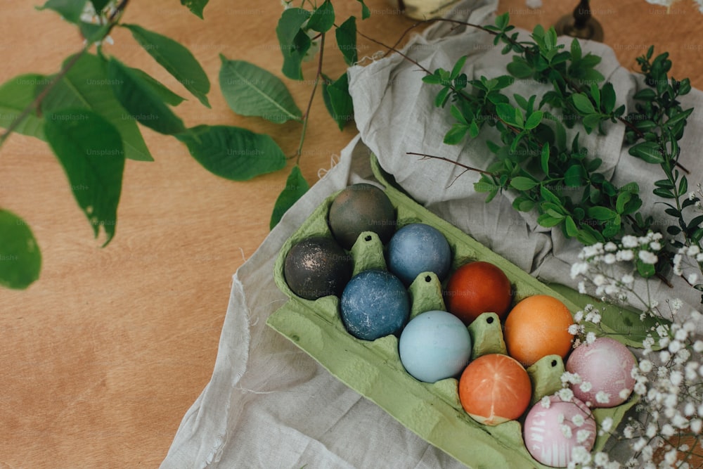 Natürliche Färbung Ostereier im Kartontablett auf rustikalem Tisch mit Blumen. Moderne gelbe, rosa, blaue und graue Ostereier bemalt mit Bio-Zwiebeln, Rüben, Rotkohl, Carcade-Tee. Zero Waste Urlaub