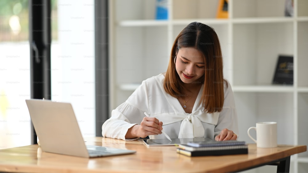 Frau hält einen Stift und zeichnet damit auf ihrem Computer-Tablet, während sie vor ihrem Laptop am modernen Arbeitstisch mit Büroglaswand als Hintergrund sitzt.