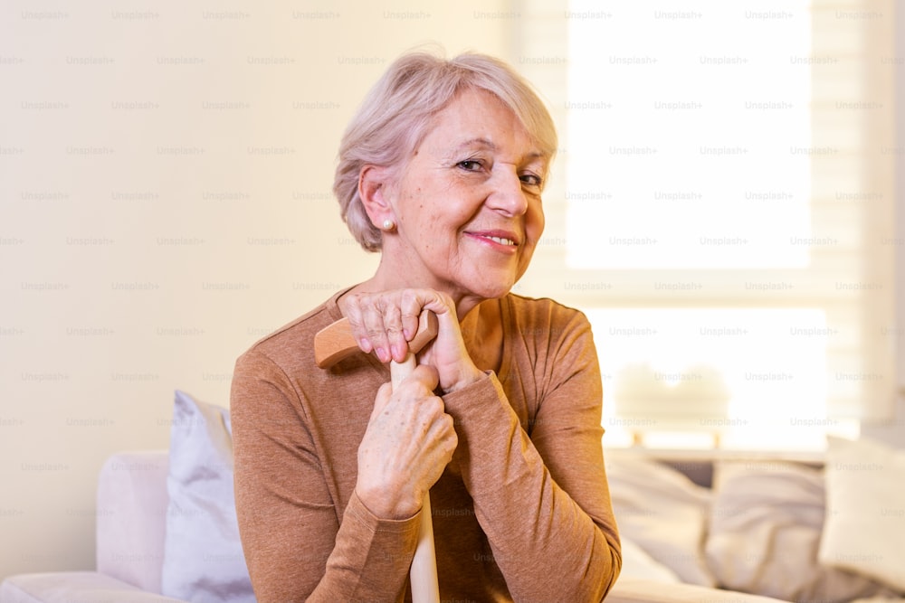 Portrait of beautiful senior woman with white hair and walking stick. Portrait of senior woman sitting on sofa at home. Smiling middle aged mature grey haired woman looking at camera