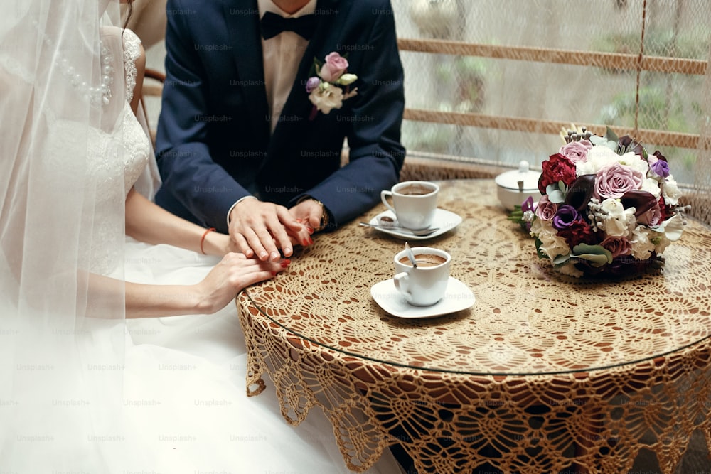 stylish wedding couple holding hands, sitting at wooden vintage table with warm coffee in cups in cafe. happy  bride and groom in dress and suit  hugging
