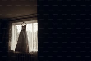 朝の光の中で窓のハンガーにぶら下がっている美しいウェディングドレス。