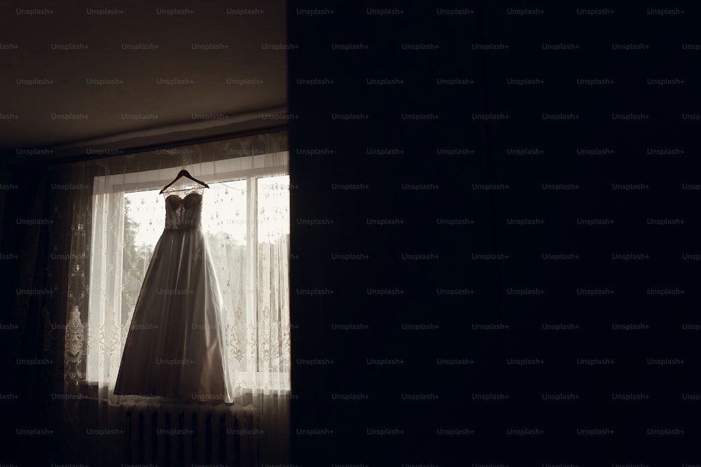 beautiful wedding dress hanging on hanger on window in morning light.