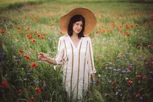 素朴なリネンのドレスを着た若い女性が、夕暮れの光の中、夏の牧草地の田園地帯でポピーとヤグルマギクの間を歩いています。田舎のスローライフ。シンプルな生活を楽しむ。野の花の帽子をかぶった女の子