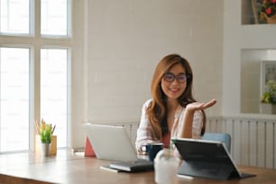 Mujer joven y hermosa con gafas que trabaja como contadora sonriendo y trabajando/sentada en la mesa de trabajo moderna con una cómoda sala de estar como fondo.