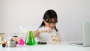 Photo of adorable schoolgirl doing a scientific experiment at the modern white table with chemistry glassware over the white laboratory wall as background. Education for kids concept.