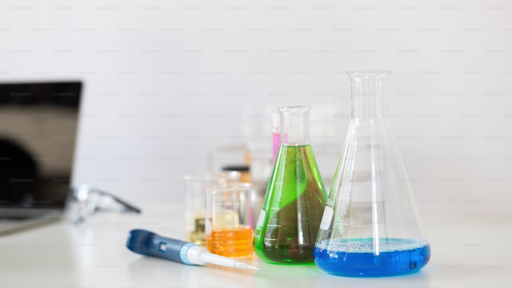 Photo of laboratory glassware containing colored liquids, Computer laptop and safety glasses while putting together on white table isolated over white background. Scientific experiment concept.