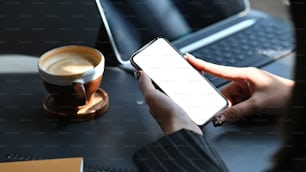 Imagen recortada de las manos de una mujer ejecutiva sosteniendo / usando un teléfono inteligente negro recortado con una pantalla blanca en blanco con una computadora portátil y una taza de café juntas en la mesa como fondo.
