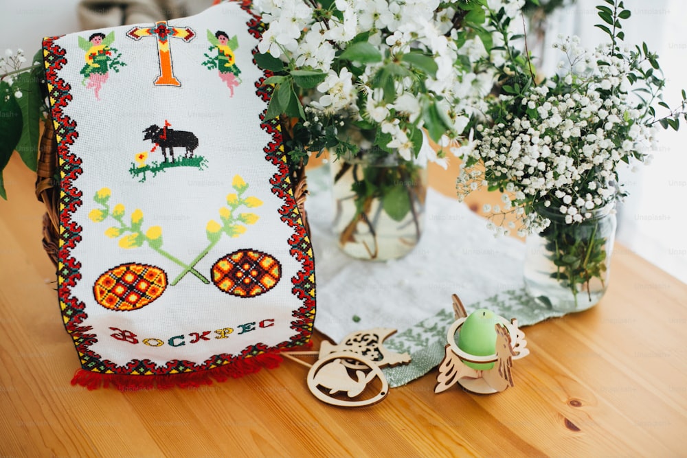 Traditionelles ukrainisches Ostersticktuch mit Eiern, Kreuz und kyrillischem Zeichen Christus ist auferstanden, bedeckter Korb mit Essen zum Segen auf Holztisch mit Kerze, grünen Zweigen und Blumen