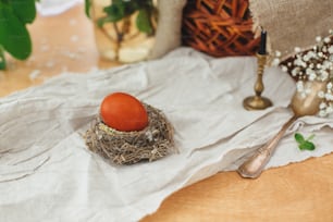 Huevo de Pascua moderno en nido en mesa rústica con flores de primavera, vela, canasta. Elegante huevo de Pascua rojo pintado en tinte natural de cebolla. Felices Pascuas
