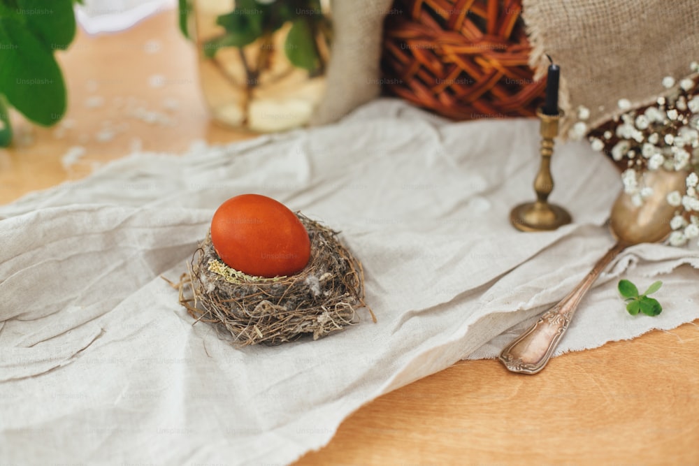 Modern Easter egg in nest on rustic table with spring flowers, candle,basket. Stylish red Easter egg painted in natural dye from onion. Happy Easter