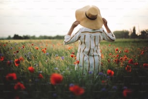 素朴なリネンのドレスを着たスタイリッシュな女性が、夕暮れの光の中でポピーや野生の花に囲まれて夏の牧草地を歩いています。雰囲気のある本物の瞬間。スペースをコピーします。田舎の帽子をかぶった女の子。田舎のスローライフ。