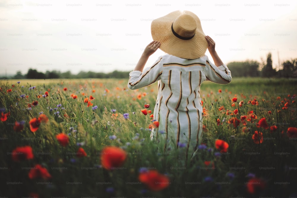 Mulher elegante no vestido de linho rústico andando no prado de verão entre papoula e flores silvestres na luz do pôr do sol. Momento autêntico atmosférico. Espaço de cópia. Menina no chapéu no campo. Vida rural lenta.