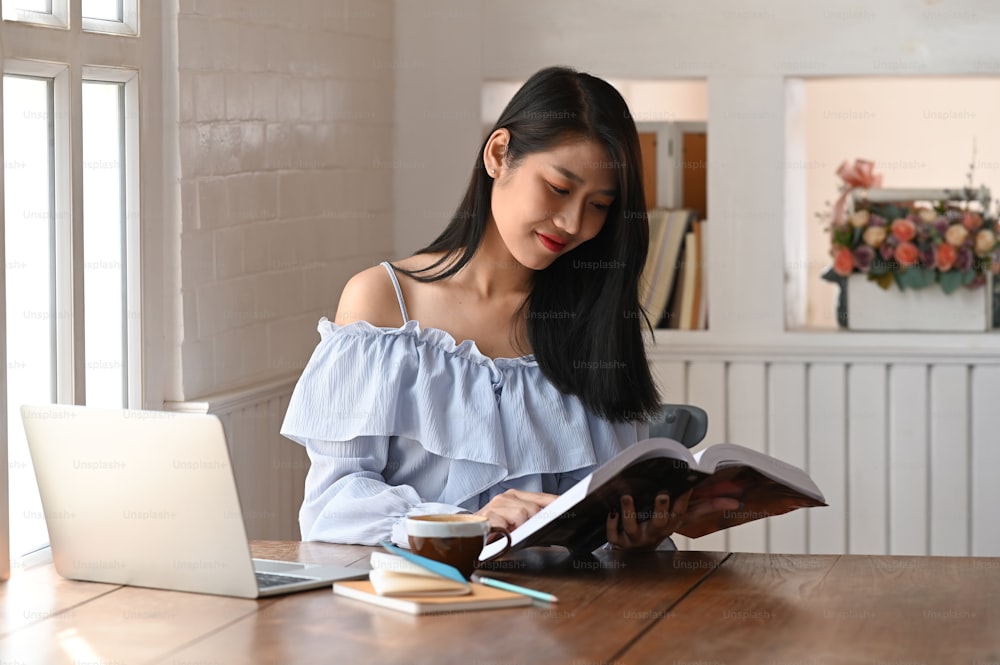 A beautiful woman reading a book while sitting at modern wooden desk.