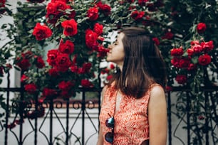 beautiful stylish hipster woman smelling wonderful red roses in sunny street in summer. beauty of blooming in spring. space for text. atmospheric moment. happy young girl