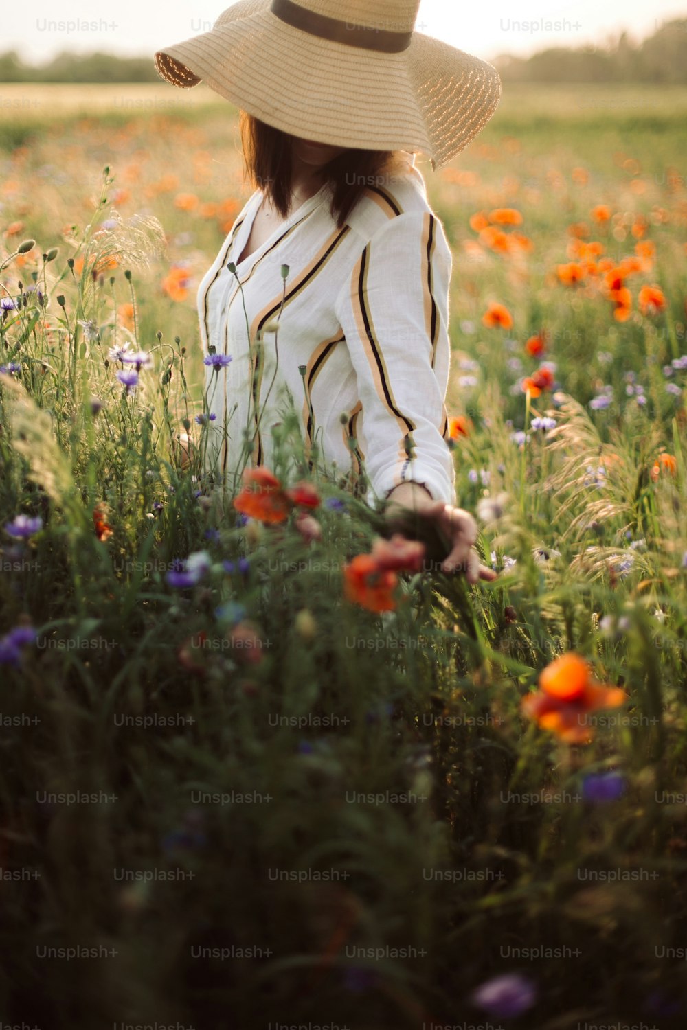 Stylish girl in hat walking in wildflowers in sunset light in summer meadow. Young woman in linen dress walking among poppy and cornflowers in countryside. Rural slow life. Enjoying simple life