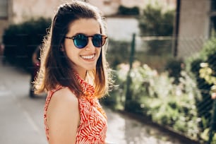 stylish hipster woman smiling in sunglasses and enjoying sunshine in sunny street in summer. young girl posing and laughing. space for text. joyful moment. summer travel