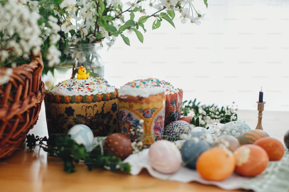 Traditional Easter food on rustic table. Homemade easter cake, easter eggs natural dyed, candle, green branches and flowers with wicker basket on wooden background. Easter Food for sanctify