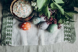 easter cake and colorful decorated eggs on rustic background with lilac flowers.  egg with floral and chicken ornaments. space for text. happy easter. greeting card concept. top view