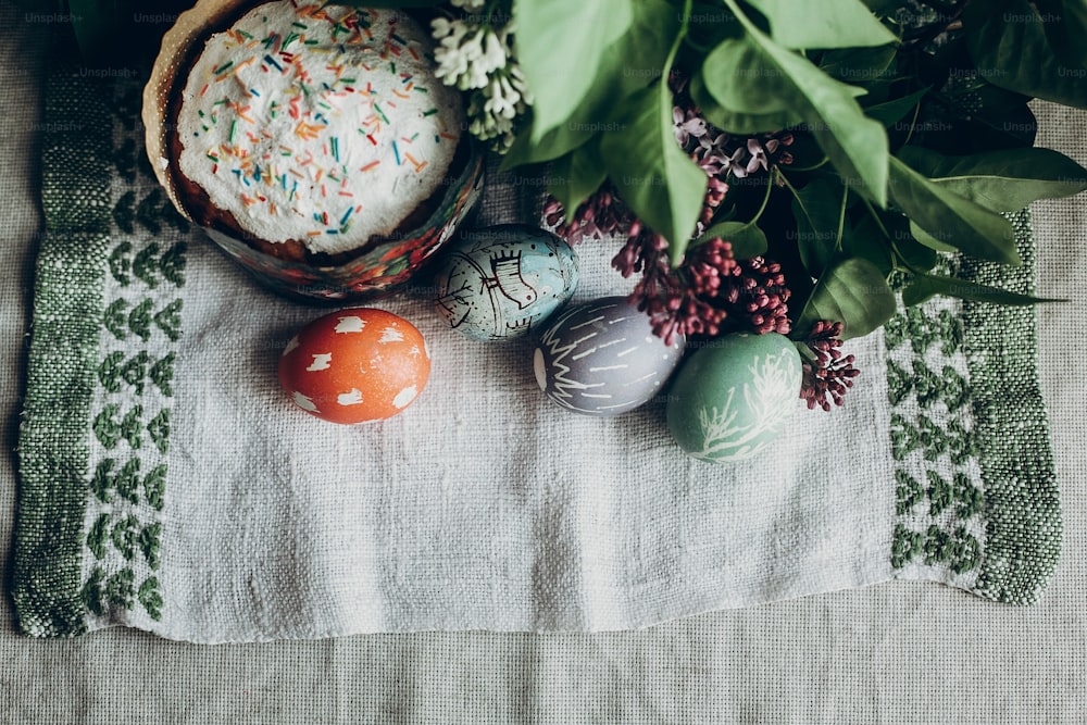 easter cake and colorful decorated eggs on rustic background with lilac flowers.  egg with floral and chicken ornaments. space for text. happy easter. greeting card concept. top view