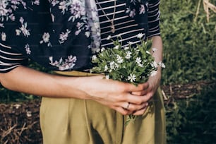 stylish hipster woman holding spring bouquet of wildflowers in sunny countryside. space for text. beautiful moment. save environment. rustic young girl with simple flowers in hand