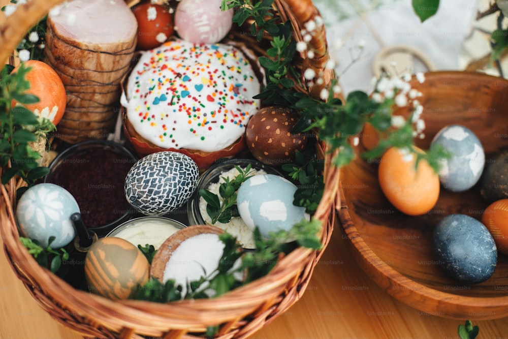 Easter modern eggs, easter bread, ham, beets, butter, in wicker basket decorated with green buxus branches and flowers on rustic wooden table. Traditional Easter basket.