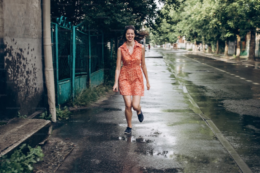 stylish hipster woman smiling and running in sunny rainy street in summer under rain with big drops. young girl enjoying moment. space for text. joyful moments. summer relax