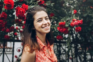 beautiful stylish hipster woman smiling under big red roses in sunny street in summer. beauty of blooming in spring. space for text. atmospheric moment. happy young girl