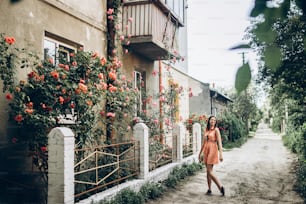 Femme hipster élégante debout à de belles roses roses et rouges sur une clôture blanche à une vieille maison dans la campagne de la rue. Provence. allée florale. printemps. espace pour le texte