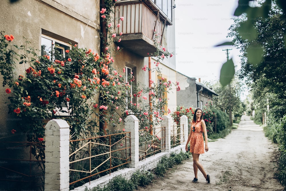 stylish hipster woman standing at beautiful pink and red roses on white fence at old house in street countryside. provence. floral alley. springtime. space for text