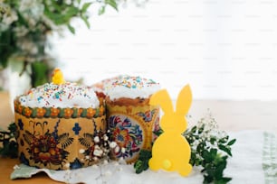 Homemade easter cake, bunny decor, green branches and flowers on wooden background. Happy holiday. Delicious easter bread with icing, colorful sprinkles and topping