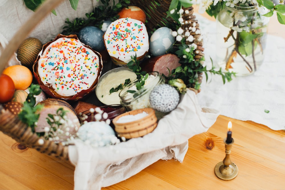 Traditional Easter basket. Easter modern eggs, easter bread, ham, beets, butter, sausages in rustic basket decorated with green boxwood branches and flowers on wooden table with candle